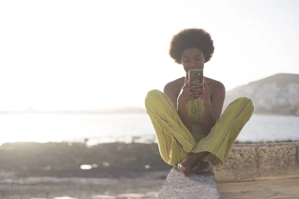 Młoda Kobieta Afrykańskich Siedząc Brzegu Przy Użyciu Smartfona — Zdjęcie stockowe