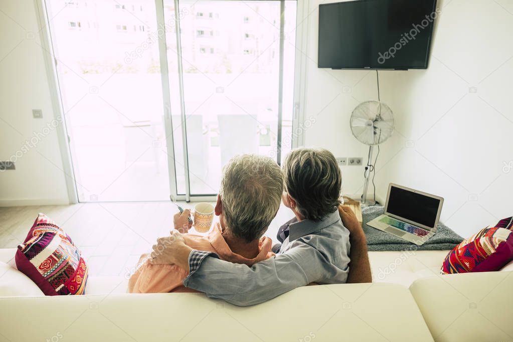 rear view of senior couple sitting at home on white sofa and drinking tea