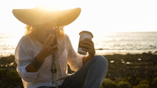 Γυναίκα Καπέλο Κάθεται Και Χρησιμοποιώντας Smartphone Φόντο Ηλιοβασίλεμα — Αρχείο Βίντεο