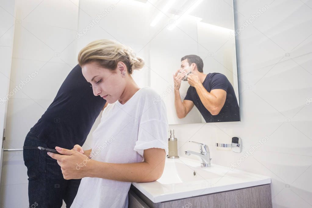 blonde young woman looking at smartphone while man doing beard with foam at bathroom 