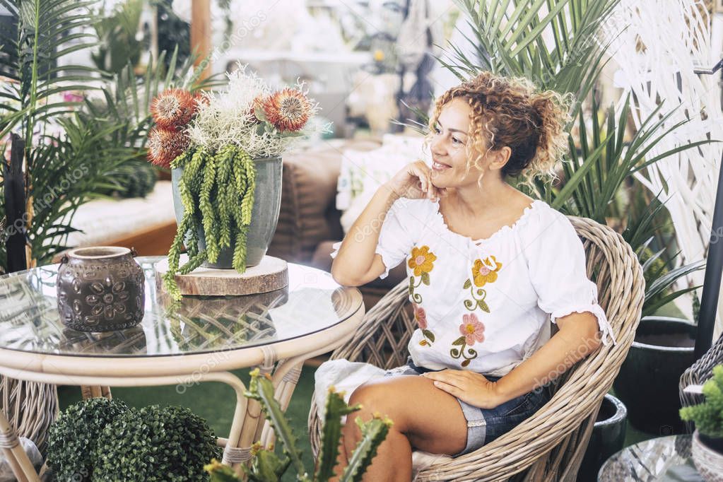 Young woman with blond curly hair sitting on a wicker chair with many plants admires the vegetation