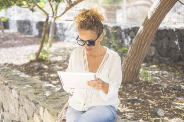 Parkta çalışan orta yaşlı güzel bir kadın internet ve dokunmatik ekran cihazı kullanan modern bir tabletle çalışıyor. Özgür insanlar için bir dizüstü bilgisayar konseptinde açık ofis dijital göçebe
