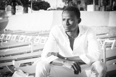 Moda siyah-beyaz resim. Afrikalı güzel model adam sahilde oturuyor. Önüne bakıyor. 