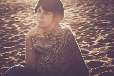 Orta yaşlı genç bir kadının romantik portresi. Sahilde oturuyor. Arka planda güneş ve günbatımı var.