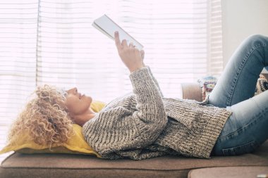 Güzel orta yaşlı kadın, evde kanepede uzanıp kitap okuyor ve dışarıdaki ışığın tadını çıkarıyor.