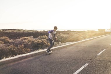 Aktif genç bir çocuk kaykay aktivitesi yapıyor. Kaykay kaykayıyla uzun bir asfalt yolda. Arka planda gün batımı ışığı var.