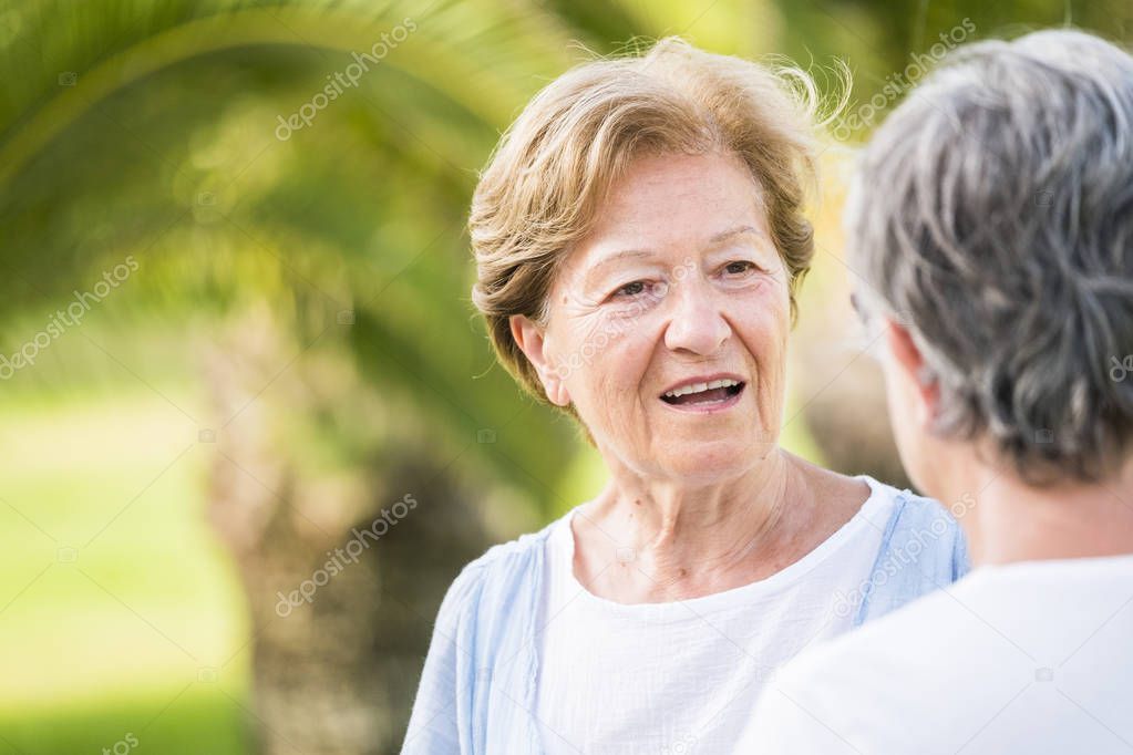 Couple of senior adult friends females talk together in outdoor leisure activity - retired lifestyle for silver society and beautiful females aged with 70 years old about - green bokeh background