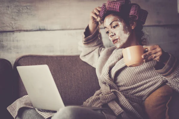 woman with facial mask and curlers hair drinking coffee while using laptop at home