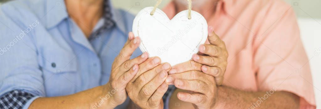close up of happy senior couple holding wooden handmade heart while sitting on sofa at home 