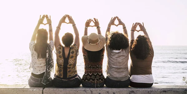 Group of young activist women viewed from rear doing feminism symbol and female power and respect with hands - sun and outdoor nature and sunset in background for group people