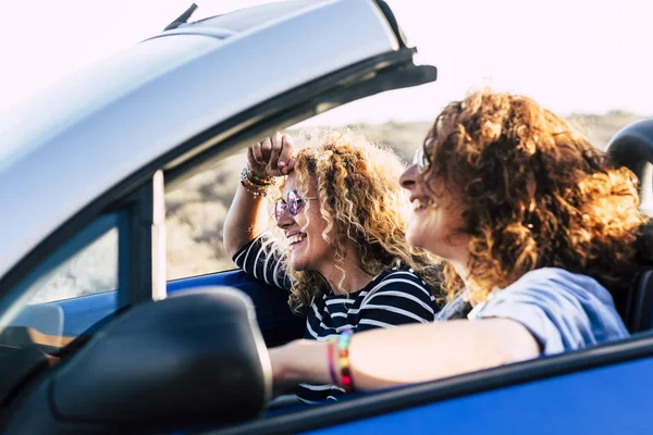 Two curly beautiful women friends drive and travel together on a convertible blue car having fun