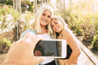 Cheerful beautiful people couple of blonde girls together enjoying the outdoor leisure activity in a sunny day taking a picture from a freind with mobile phone to share on internete social media - sun backlight