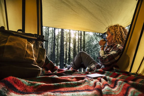 独立した美しい巻きブロンドの髪の女性はお茶を楽しみながら森の中で無料の野生のキャンプでテントの外に座って考えて — ストック写真