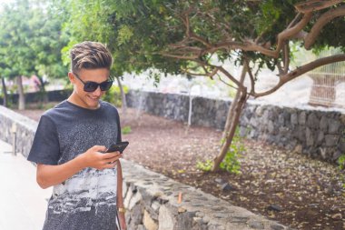 Yakışıklı genç beyaz çocuk cep telefonunu açık havada kullanıyor. 