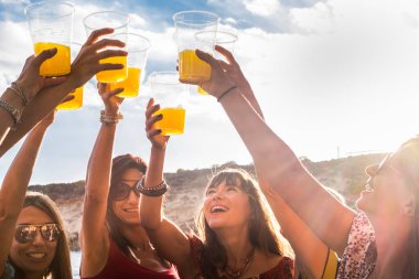 Bir grup neşeli beyaz kadın arkadaşlığın ve meyve suyuyla kadeh kaldırmanın tadını çıkarıyor. Arka planda insanları ve mavi gökyüzünü kutluyorlar.