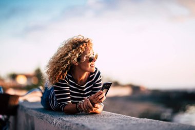 Güzel, orta yaşlı, sarışın kıvırcık kadın dışarıda eğleniyor ve dinleniyor. Günbatımını seyrediyor. Modern telefonlardan arkadaşlarını aramaya hazır. 