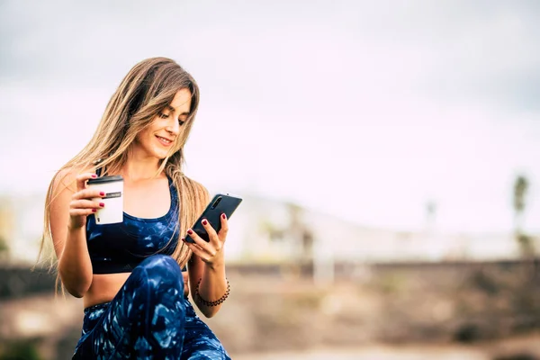 时尚双语 快乐美丽的年轻女性在户外运动后休息一下 一边喝咖啡 一边看并使用现代超市手机 — 图库照片