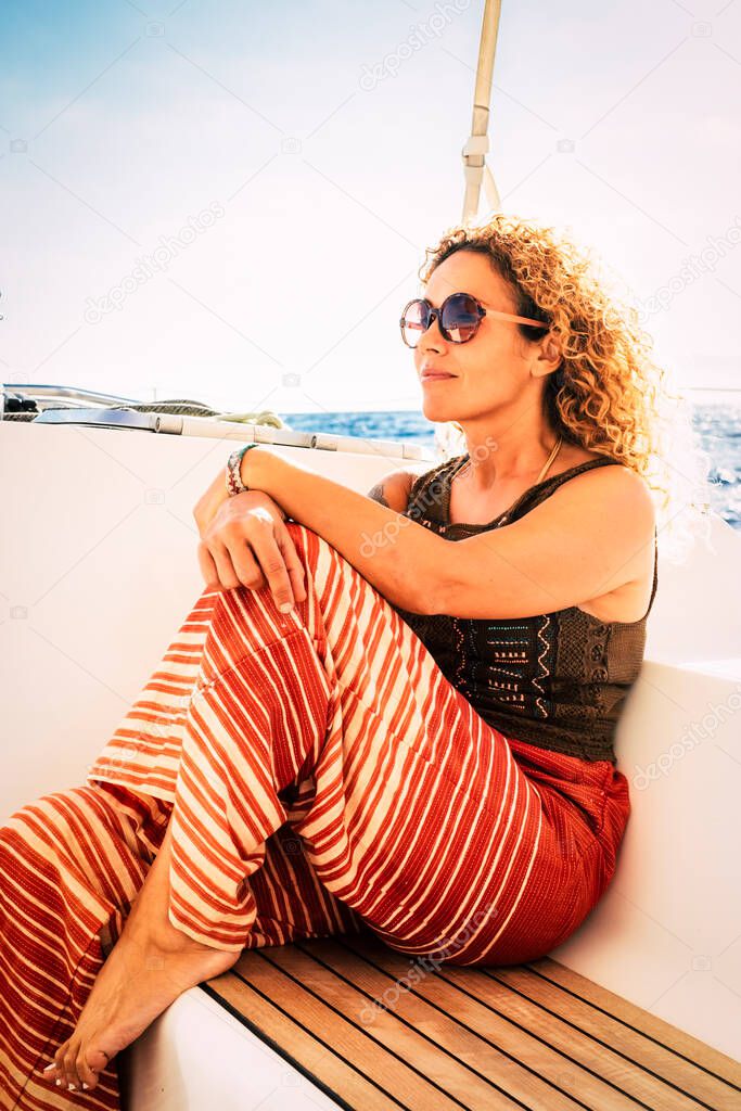 Beautiful trendy coloured clothes caucasian adult woman enjoy sailing on the boat sit down on the wood bench - yachting summer vacation activty - blue sky and ocean in background