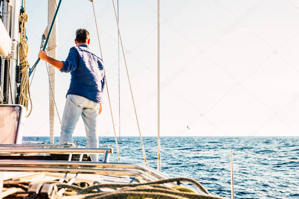 man viewed from back stand up on the deck of a sail boat looking horizon and blue ocean and sky