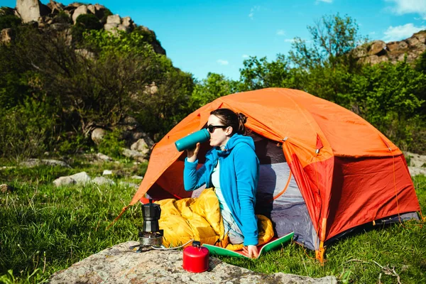 Camping in the forest. A girl is preparing coffee on a geyser coffee machine. A woman sits near a tent in a sleeping bag and prepares coffee against the background of the mountains.