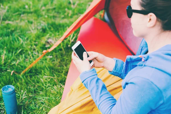女の子はテントの近くに座っています 女性が携帯電話を見て 笑みを浮かべてします 寝袋で観光 自然の中のキャンプ モバイル通信 — ストック写真