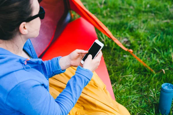 女の子はテントの近くに座っています 女性が携帯電話を見て 笑みを浮かべてします 寝袋で観光 自然の中のキャンプ モバイル通信 — ストック写真