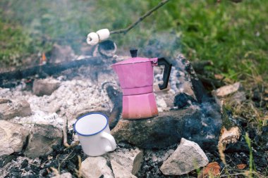 Bir kupa ve şofben kahve makinesi şenlik ateşi yakın. Ormanda kamp. Bir fincan bir yangın çerçevede çim üzerinde duruyor. Söz konusu Kavrulmuş Lokum.