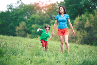 Çocuk annesiyle çayır yürür. Bir kadın oğlunu elinden tutar. Çocuk ve anne alan yürüyor. Yeşil çimen.