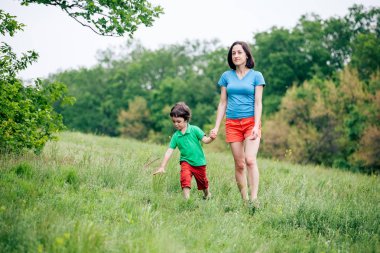 Çocuk annesiyle çayır yürür. Bir kadın oğlunu elinden tutar. Çocuk ve anne alan yürüyor. Yeşil çimen.