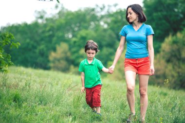 Çocuk annesiyle çayır yürür. Bir kadın oğlunu elinden tutar. Çocuk ve anne alan yürüyor. Yeşil çimen.