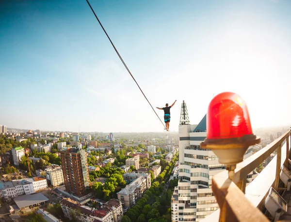 A man walks along a line stretched between two buildings. A man catches balance at altitude. Highline over the city.