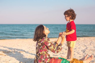 Annenin bebeği ile sahilde çalış. Oğlu deniz kıyısı üzerinde olan bir kadın. Bir çocuk ve annesi kum üzerine oturuyor.