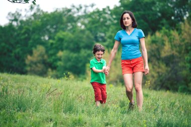 Çocuk annesiyle çayır yürür. Bir kadın oğlunu elinden tutar. Çocuk ve anne alan yürüyor. Yeşil çimen.