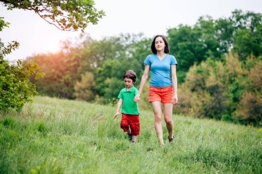 Çocuk annesiyle çayır yürür. Bir kadın oğlunu elinden tutar. Çocuk ve anne alan yürüyor. Yeşil çimen.