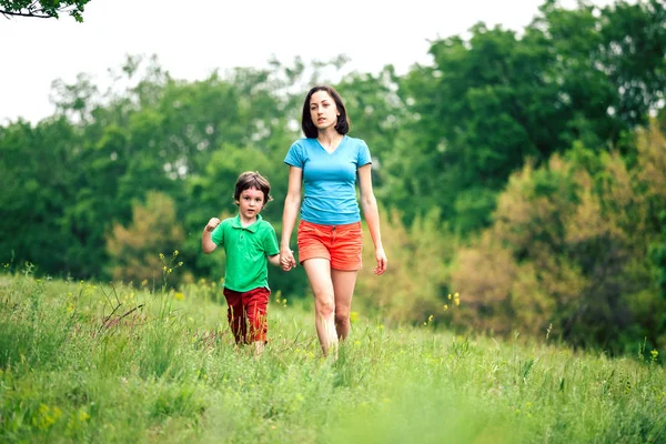 Çocuk annesiyle çayır yürür. Bir kadın oğlunu elinden tutar. Çocuk ve anne alan yürüyor. Yeşil çimen.