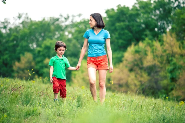 Çocuk annesiyle çayır yürür. Bir kadın oğlunu elinden tutar. Çocuk ve anne alan yürüyor. Yeşil çimen.