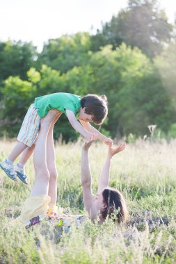Anne oğlu aktif oyunlar oynuyor. Jimnastik alanında bir çocukla. Çocuk Anne bacaklarına yalan söylüyor. Kadın bebek ayağa yükseltir. Çimenlerin üzerinde bir çift yoga. Akroyoga.