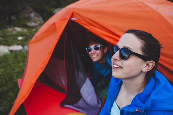 Two girls are sitting in a tent. Girlfriends communicate in nature. Women in sleeping bags drink coffee. Friends in the campsite. Smiling brunettes talking.