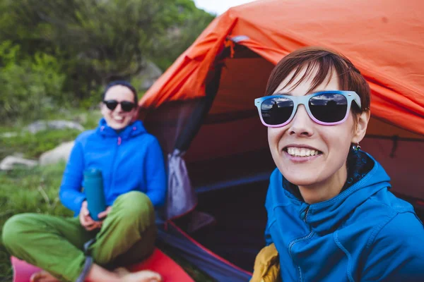 Two girls are sitting in a tent. Girlfriends communicate in nature. Women in sleeping bags drink coffee. Friends in the campsite. Smiling brunettes talking.