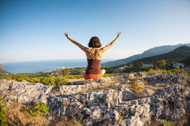 Kız dağın günbatımında oturuyor. Kadın ellerini kaldırdı. Meditasyon medeniyet çok uzak olduğunu. Güzel doğa ve tefekkür. Dağlar ve deniz görünümü.