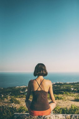 Bir kız günbatımında bir dağın tepesinde oturuyor. Bir kadın yoga yapan ve lotus pozisyonda oturuyor. Meditasyon medeniyet çok uzak olduğunu. Güzel kadın geri. Dağlar ve deniz görünümü.
