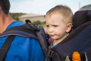 Bir çocuk bir sırt çantası içinde oturuyor. Bir çocuk portresi. Çocuk ailesiyle birlikte dolaşıyor. Bir çocukla tırmanma. İlginç çocukluk. Bir bebek taşıyan baba. Bebek gülümsüyor.