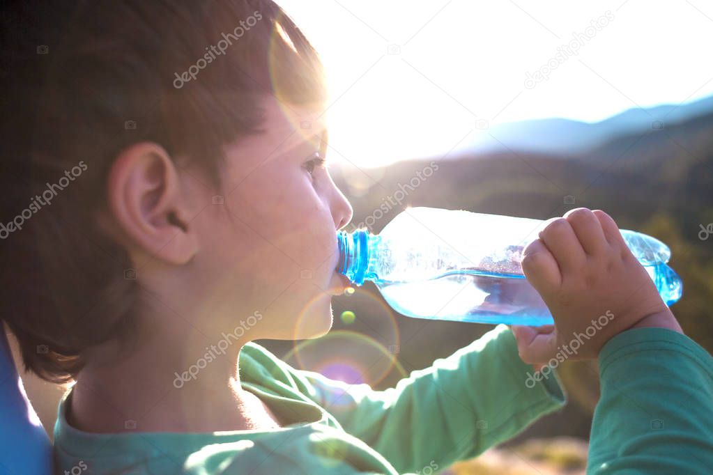 A boy drinks water from a bottle. The child quenches his thirst. The kid is traveling through the mountains. Tired child rests after a walk.