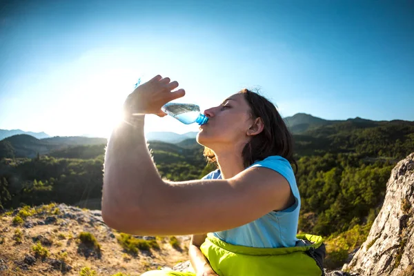 A woman is drinking water at the top of a mountain. The girl quenches his thirst. Tired tourist drinks from a plastic bottle. The brunette is sitting in a sleeping bag.