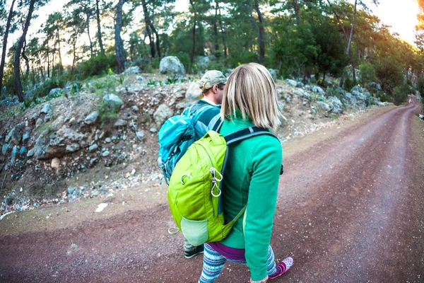 Friends go on a stony road. A woman and a man are walking in the park. A couple with backpacks travels to beautiful places. Tourists go along the mountain road.