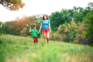 Çocuk annesiyle çayır yürür. Bir kadın oğlunu elinden tutar. Çocuk ve anne alan yürüyor. Yeşil çimen.