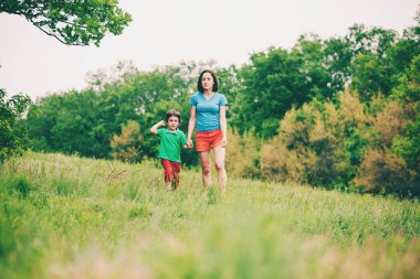 Çocuk annesiyle çayır yürür. Bir kadın oğlunu elinden tutar. Çocuk ve anne alan yürüyor. Yeşil çimen.