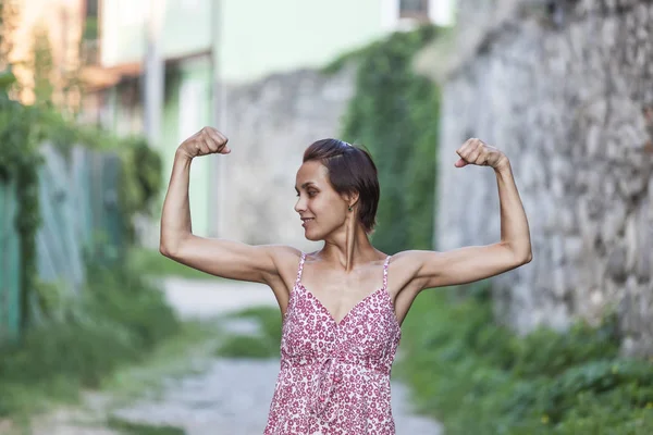 The girl shows her muscles. A woman in a summer dress shows her biceps. Strong brunette. Slender girl with strong hands.