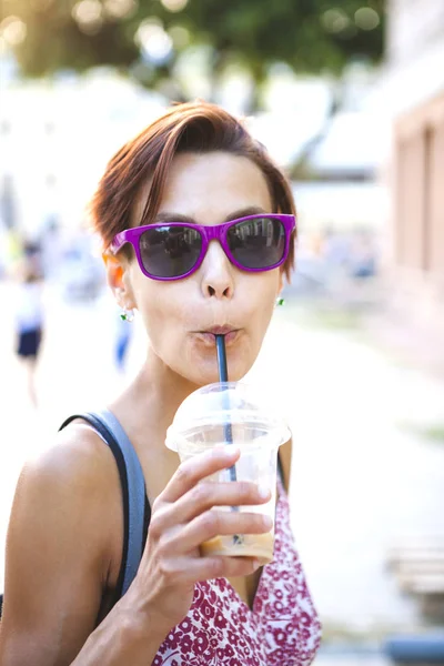 Bir kız bardaklarda şehir etrafında yürür ve sabah kahve içiyor. Gülümseyen kadın bir şehir sokak yürürken bir milkshake içmeye başlamış. Güzel bir esmer portresi yakın çekim.