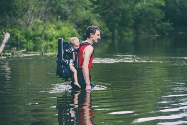 子供を持つ男は 川を横切る 息子をバックパックで運びます 洪水から救出します アジアにおける洪水犠牲者の避難 ジャングルの中を子供と一緒に旅行 極端な観光 — ストック写真